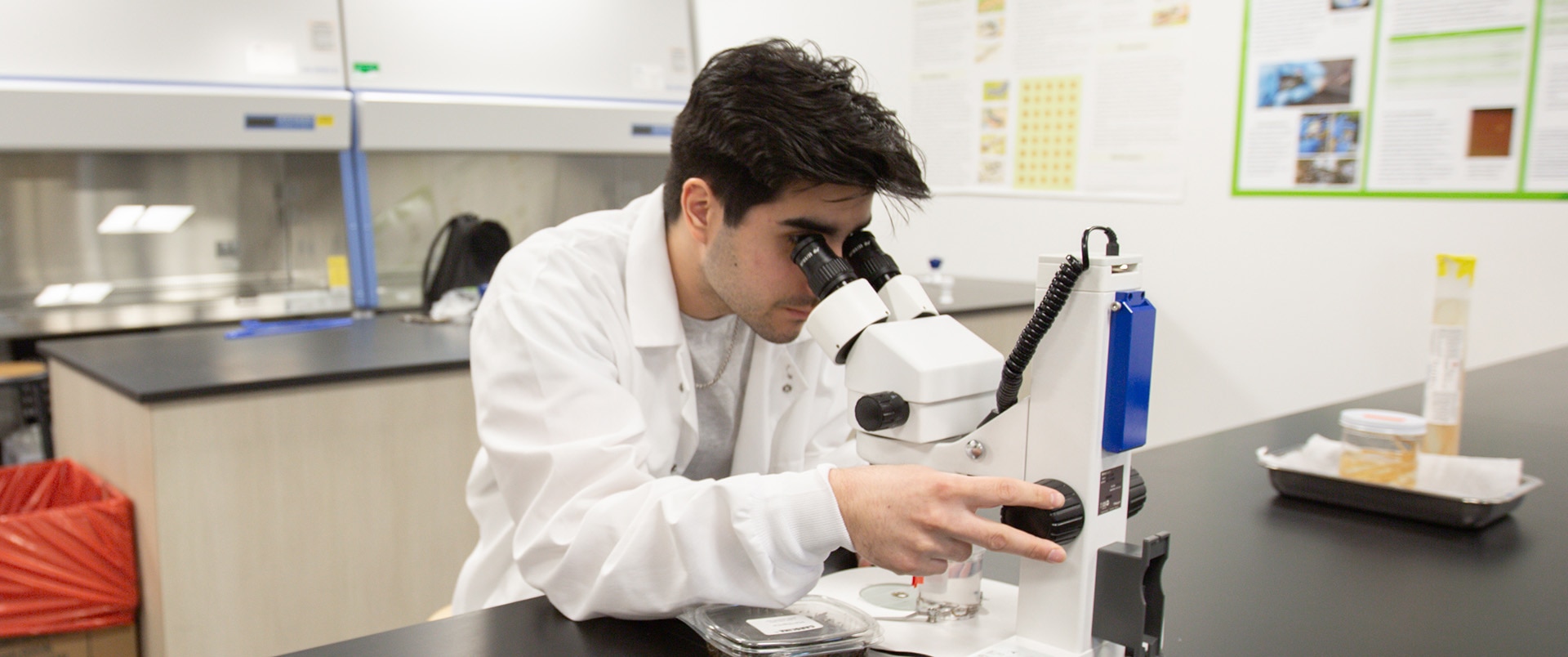 A student wearing a white lab coat looking through a microscope in a lab classroom.