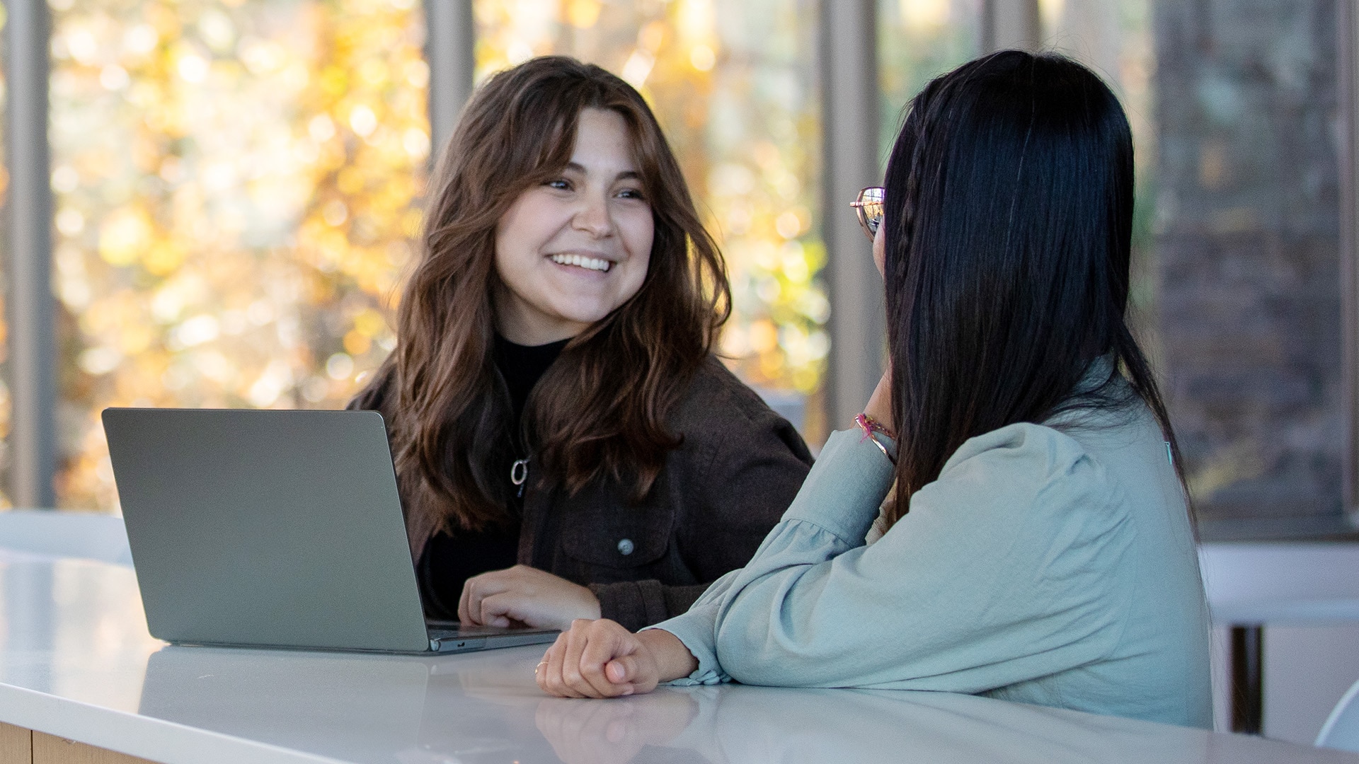 Two individuals sitting at a table, smiling and chatting, while also using a laptop.