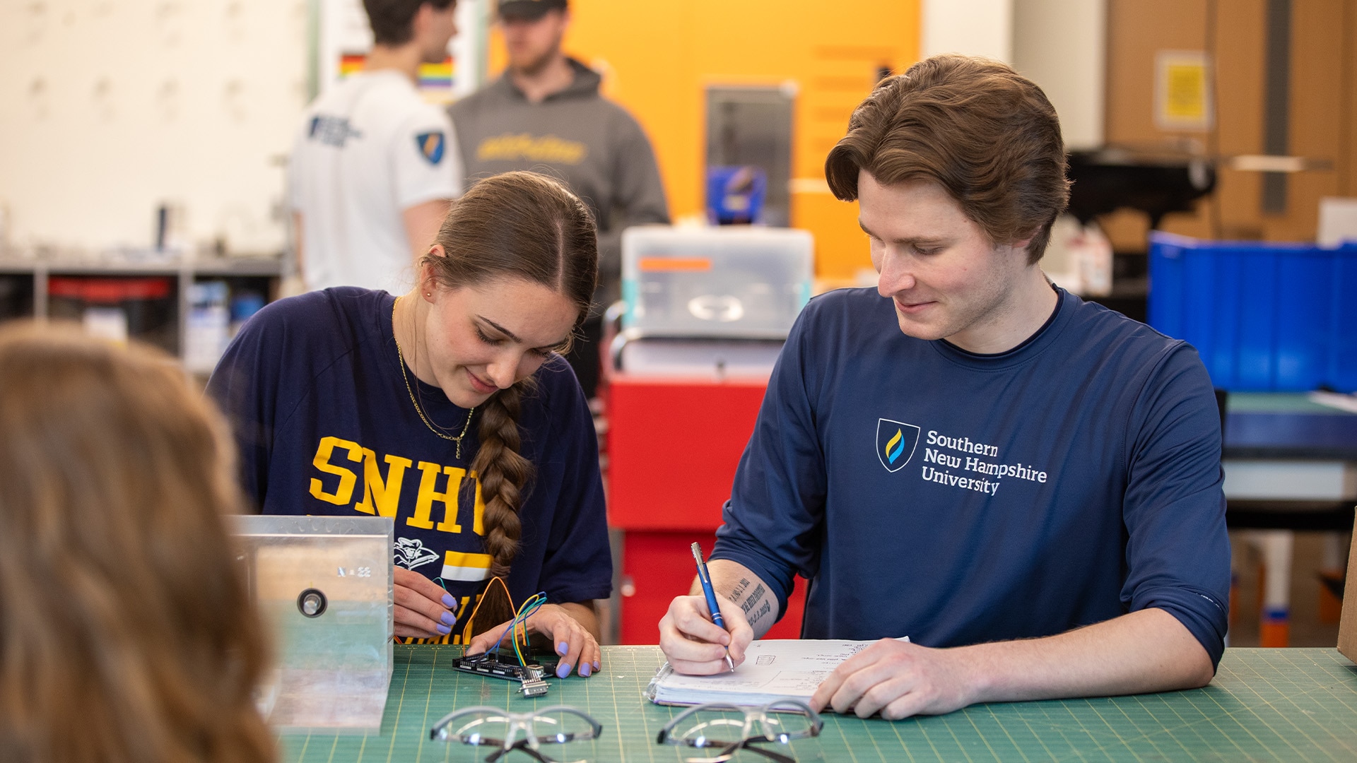 Two students wearing SNHU T-shirts working together on an electronics project at a table.