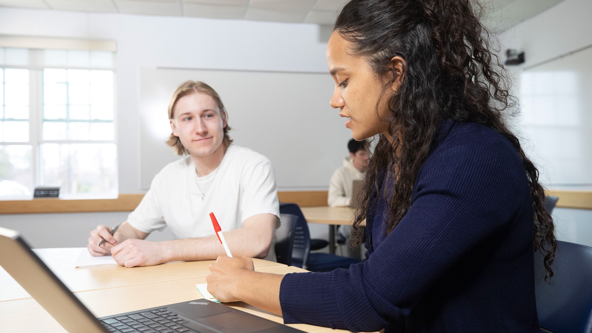 Two students discussing and making notes at a table, with a laptop in front of one of them.