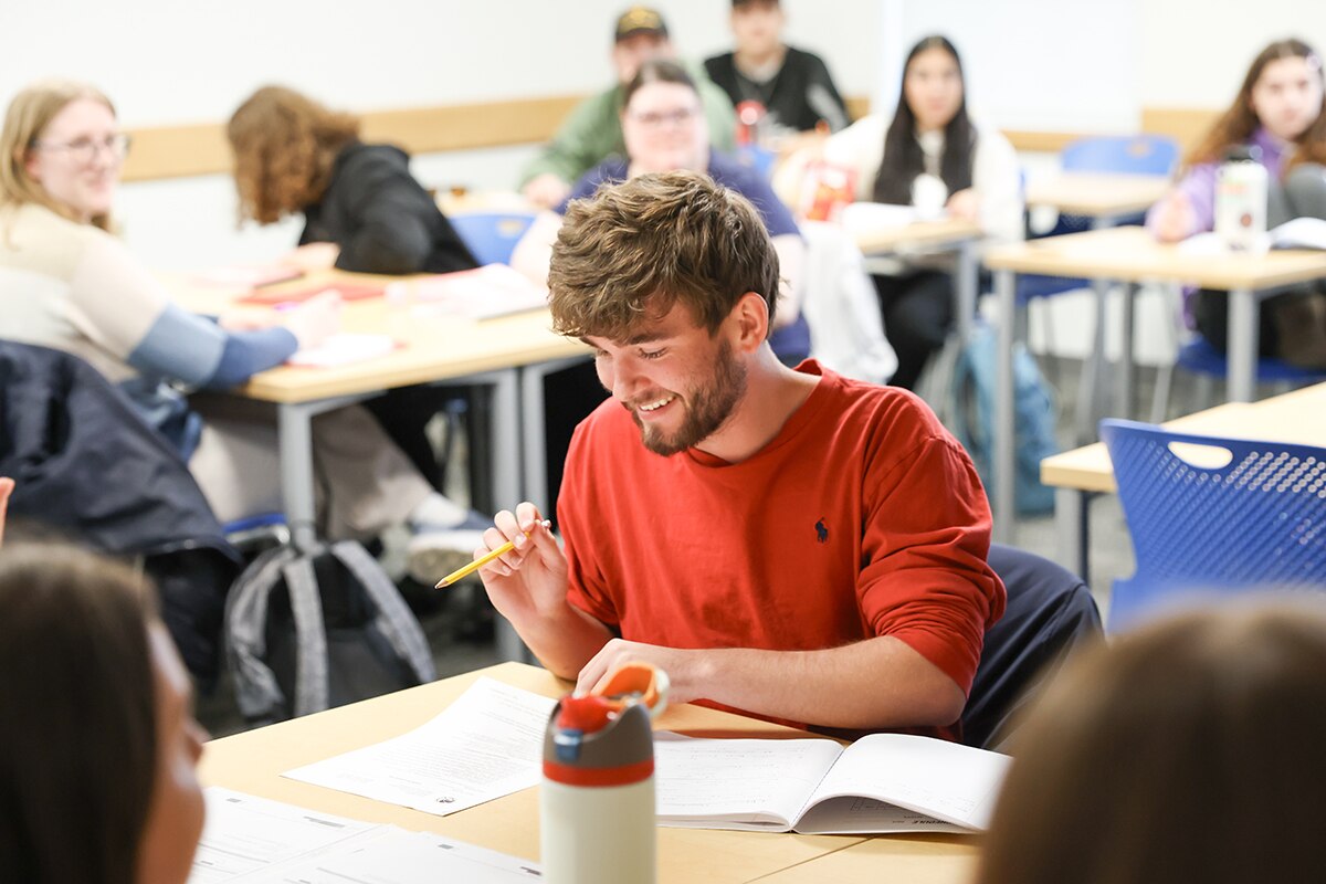A student in a classroom smiling while holding a pencil and looking at a paper on his desk.