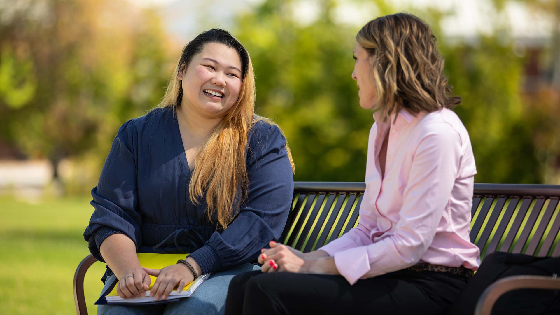 Two individuals sitting outside on a bench while talking and smiling.