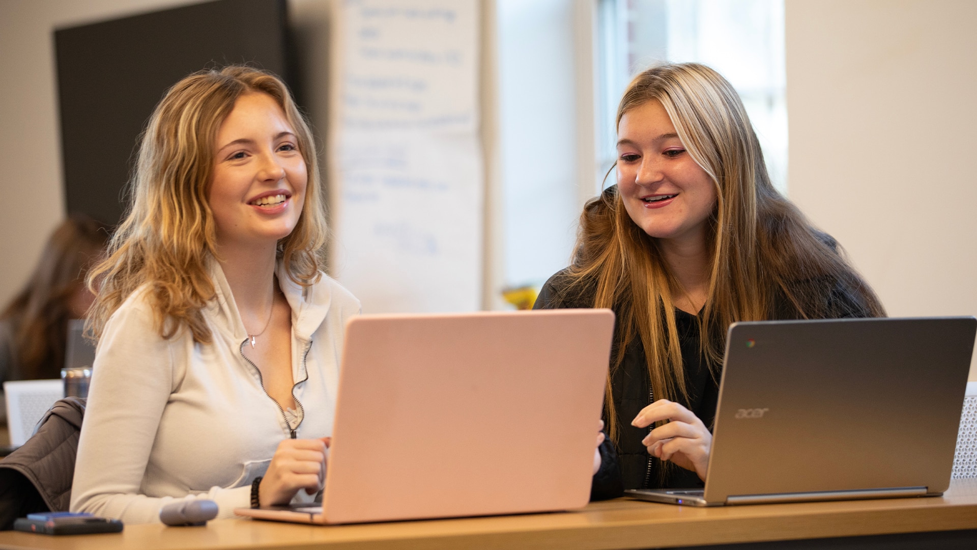 Two students smiling and discussing something while looking into their laptop screens.