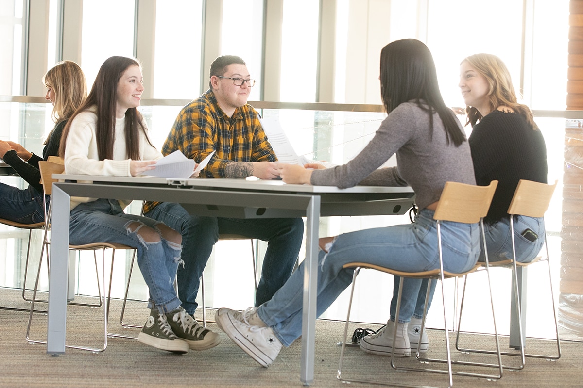 A group of 4 students sitting at a desk talking to each other while they study together.