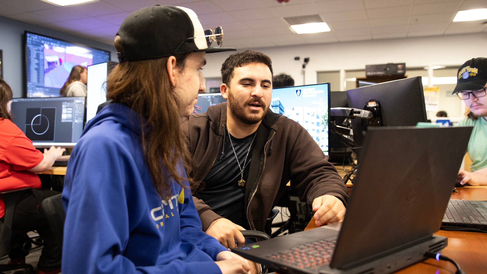 Two individuals working on a project using a laptop in a classroom setting with computers and other students.