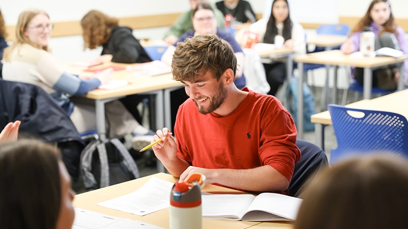 A group of students in a classroom, with one student in the foreground smiling while taking notes.