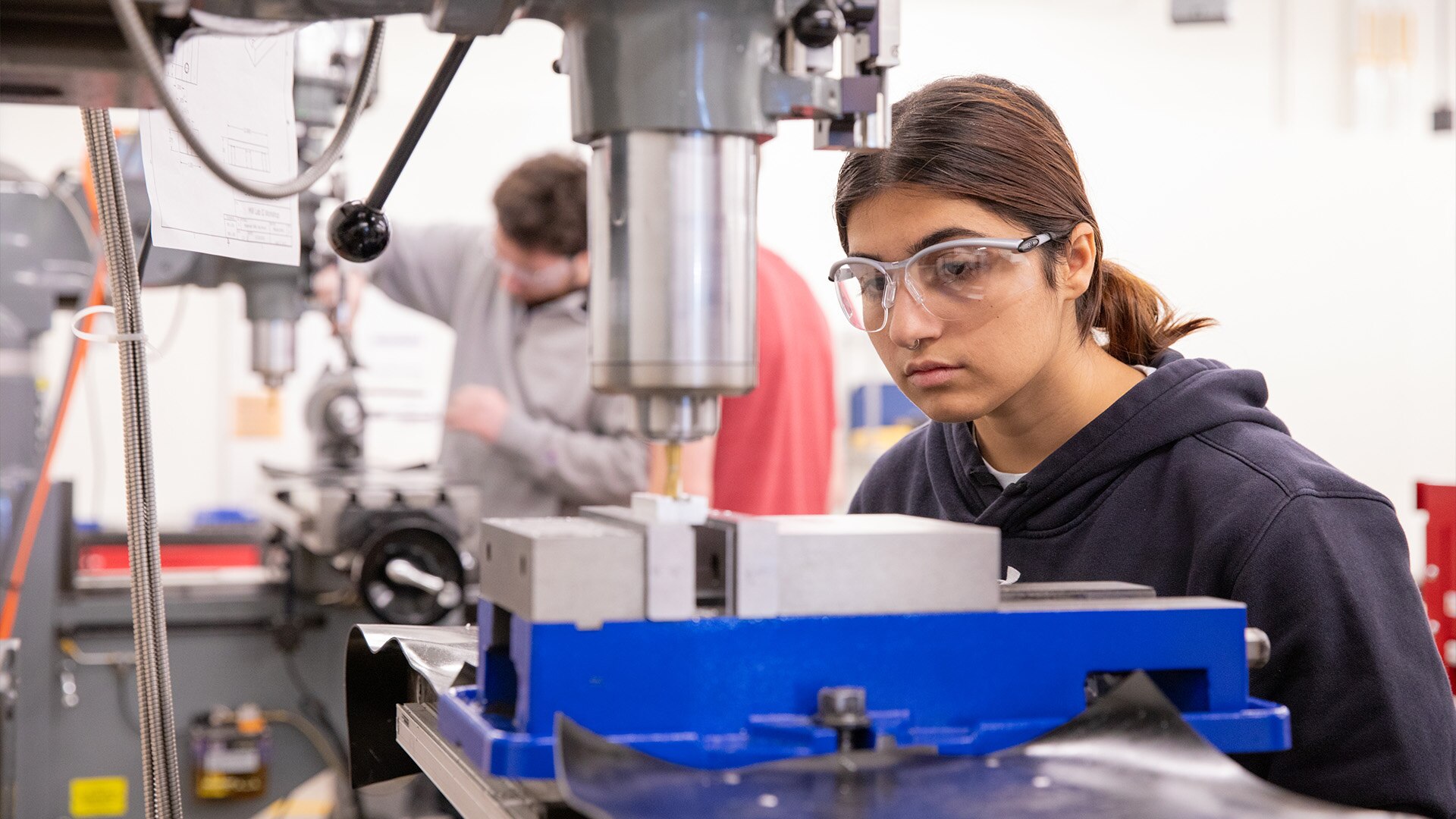 A student wearing safety glasses operates a drill press in a lab.