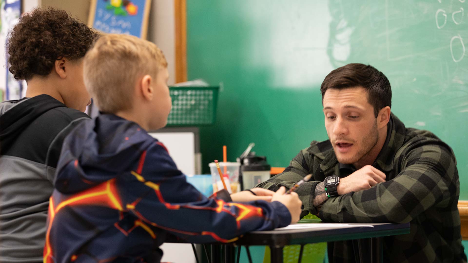 A teacher is sitting at a table, talking with two kids in a classroom.