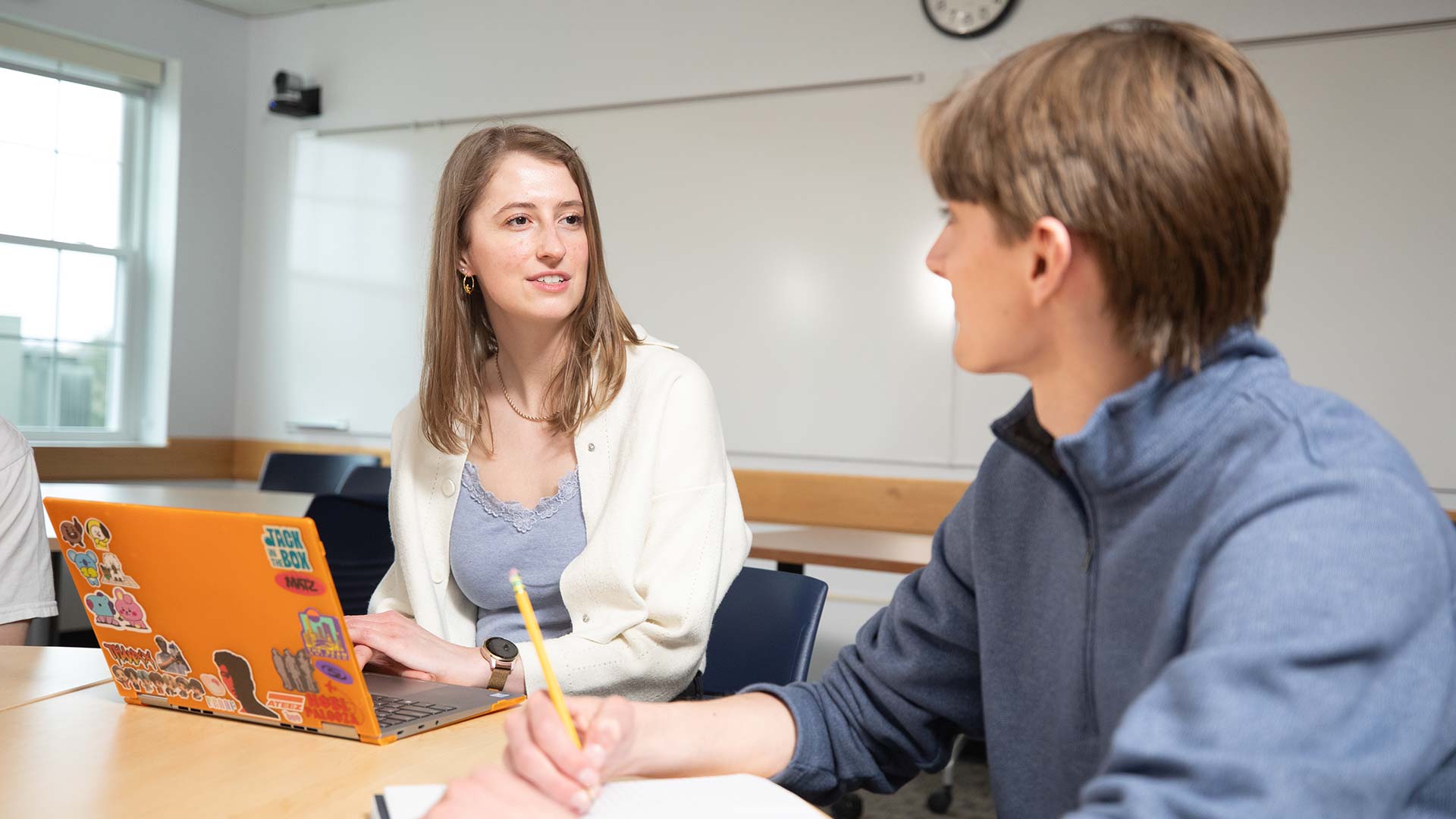 Two students sitting and discussing in a classroom, while one uses a laptop and the other uses a notebook.