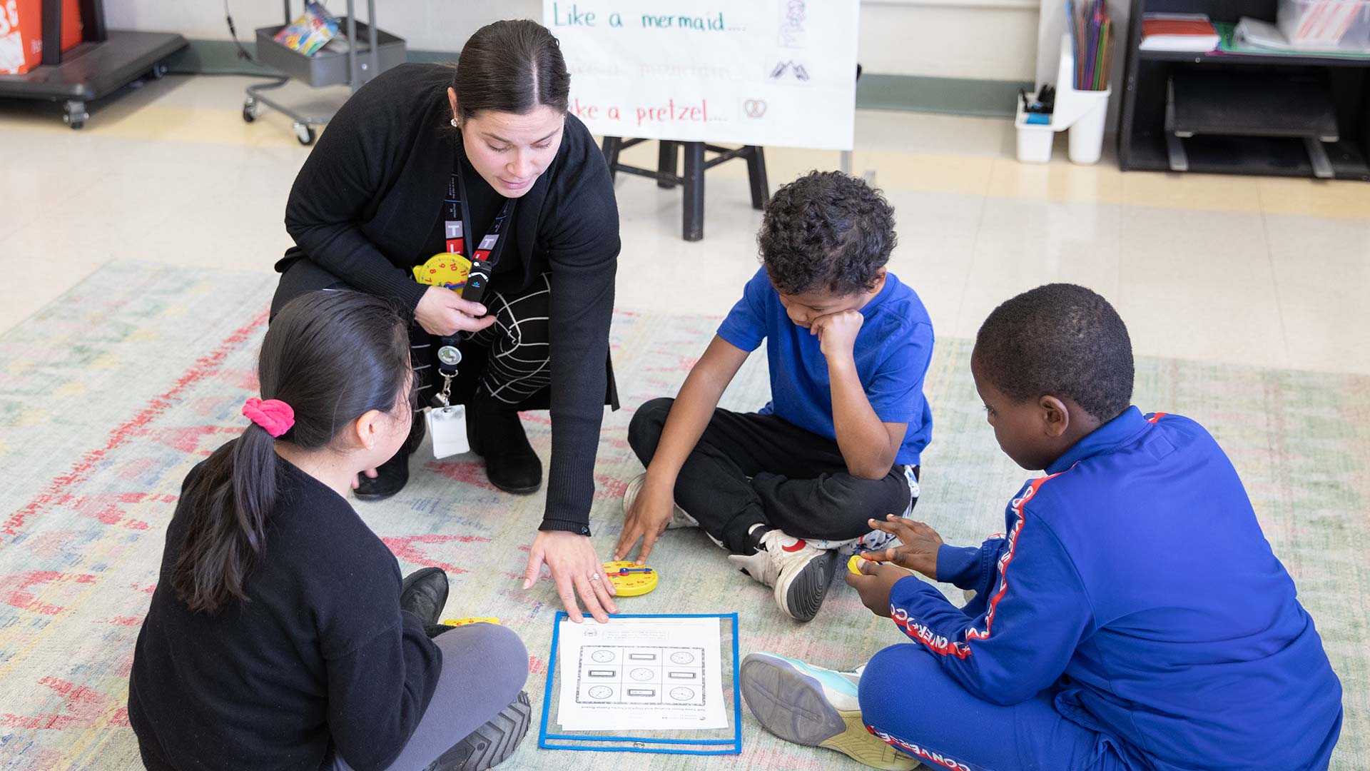 An individual teaching three kids who are sitting on the floor looking into a sheet of paper.