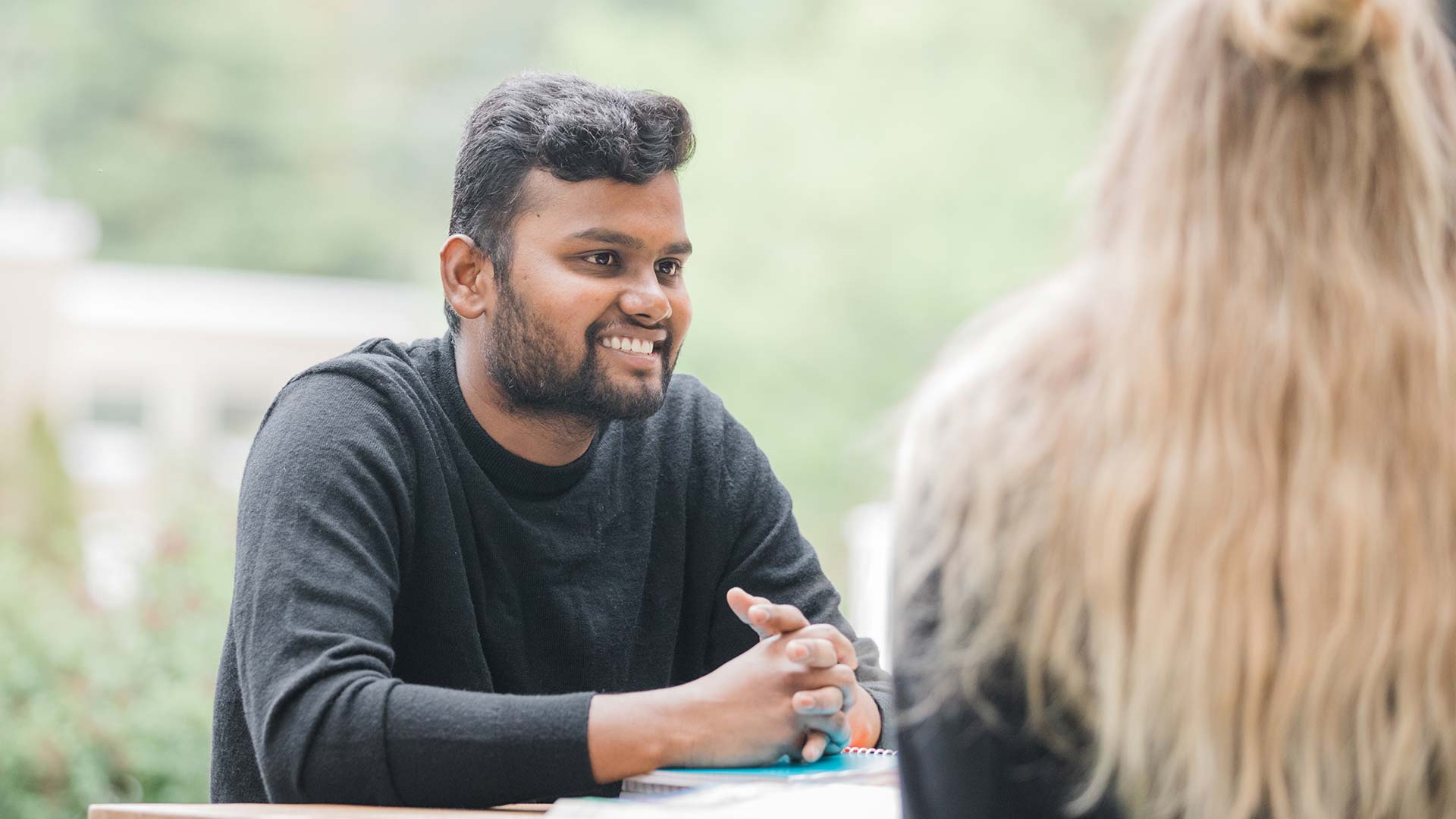 A person smiling while talking to another person outdoors.