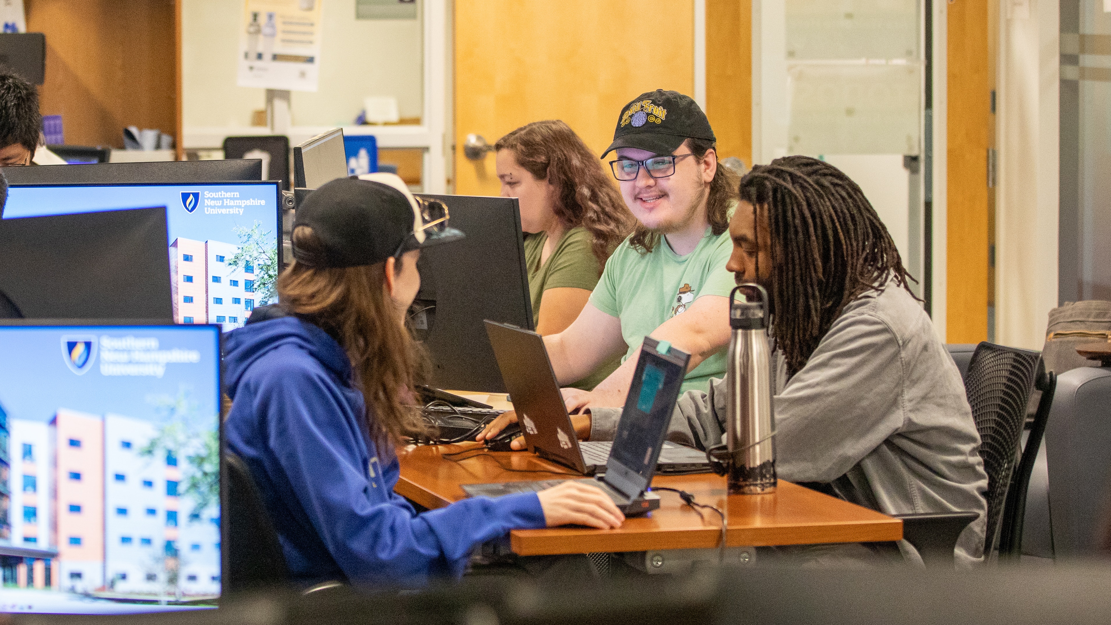 A group of students work together using laptops and desktop computers in a classroom.