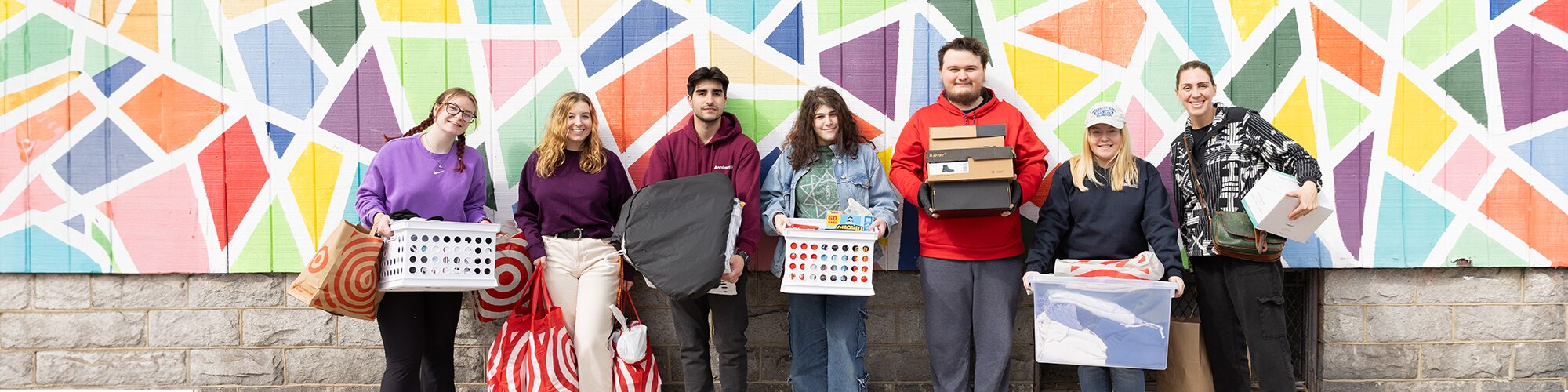 A group of seven students standing in front of a colorful geometrical mural, holding donation items for community support.