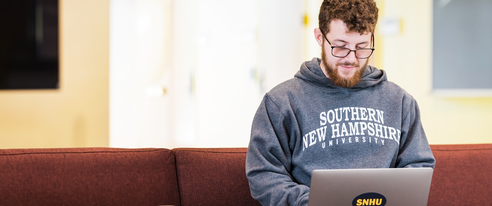 A person wearing a "Southern New Hampshire University" hoodie while using his laptop, sitting on a couch.
