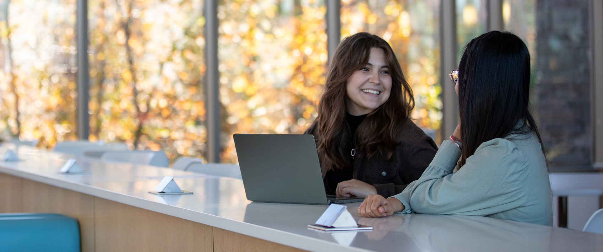 Two female students sitting in a study lounge while talking and smiling at each other.