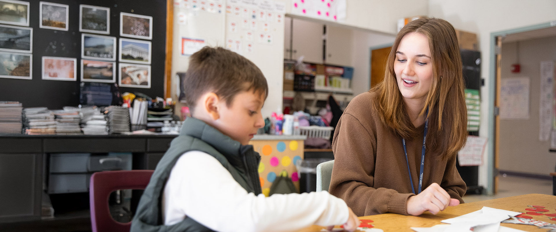 An education student and a child sitting and talking at a desk in an elementary school classroom.