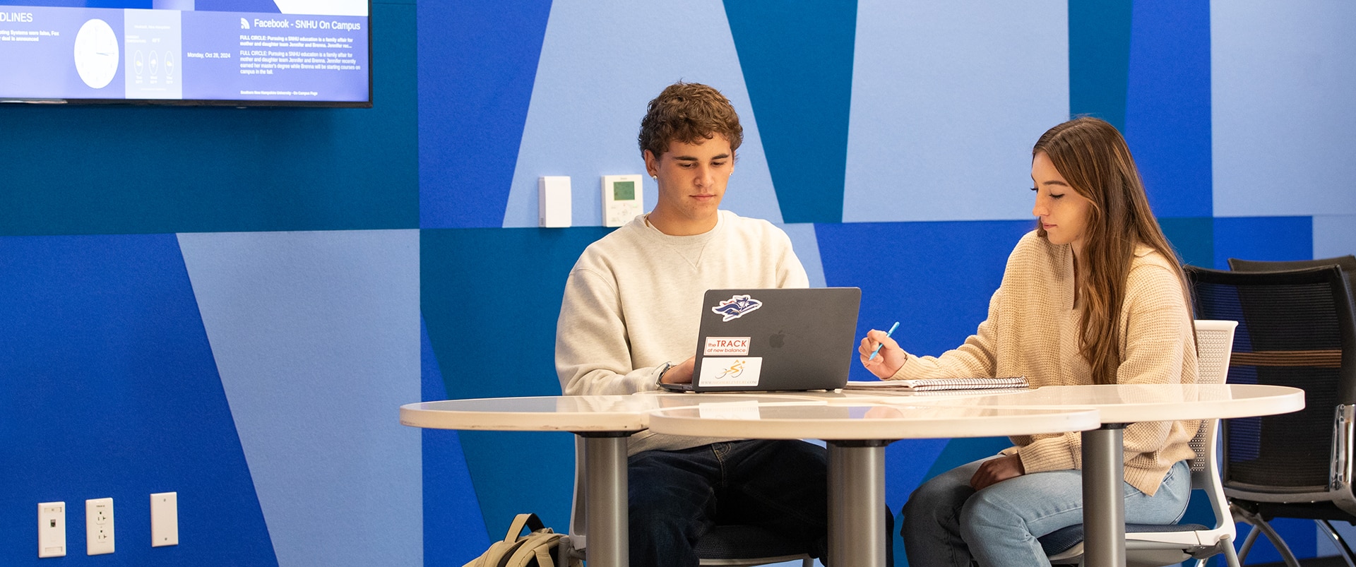 Two students sitting at a table, working together with a laptop and notebook.