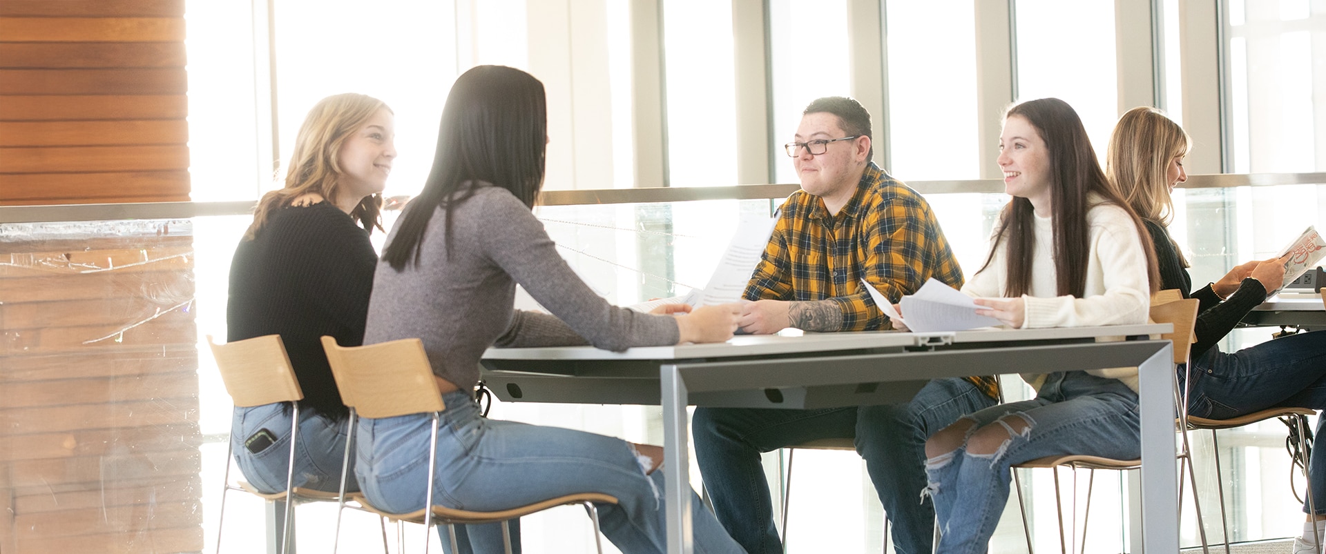 A group of four students sitting at a desk in a study lounge while holding papers and talking to each other.