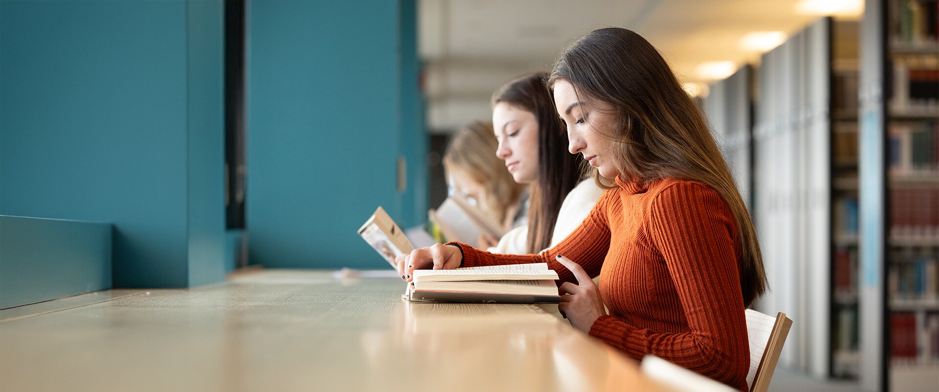 Three students sitting at a desk, each reading a book in a library.
