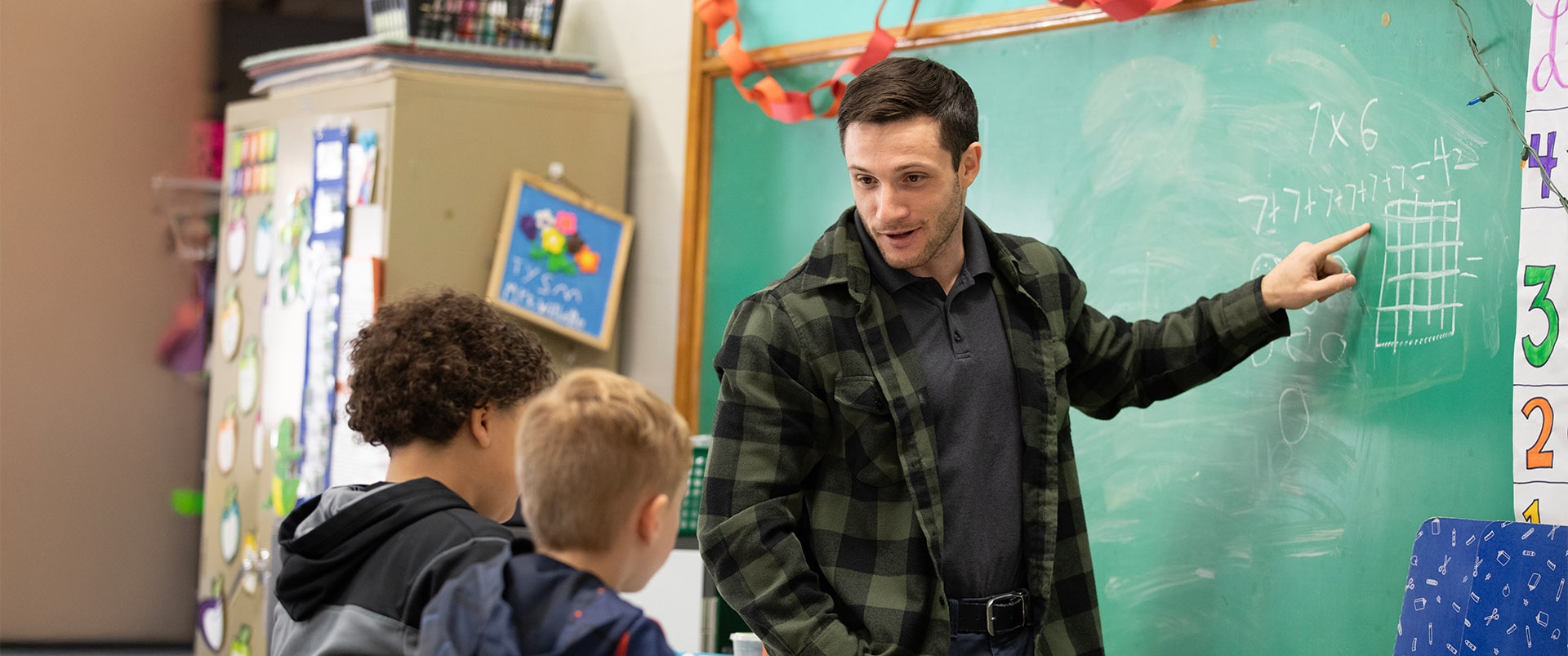 An instructor in a classroom pointing to a figure on a chalkboard while speaking to two children.