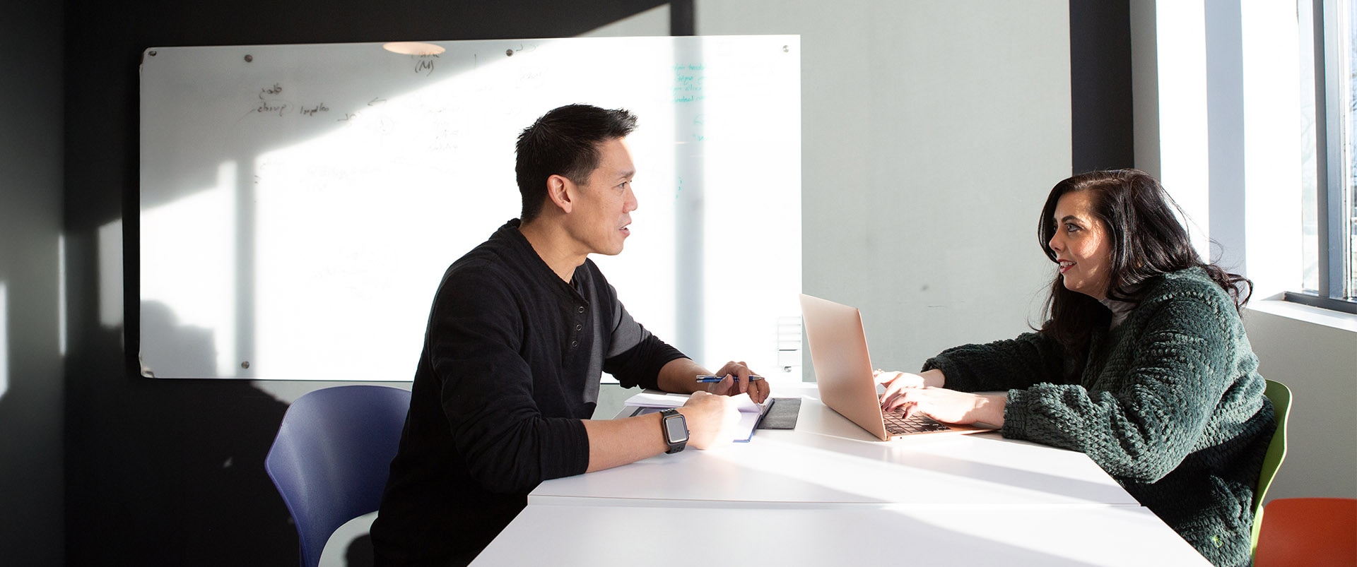 A student and an instructor talking to each other at a desk while the student uses a laptop.