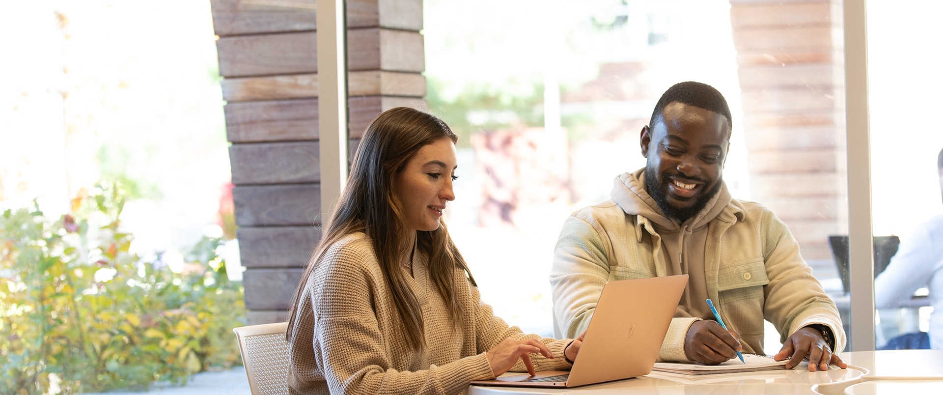 Two individuals sitting at a table, working together on a laptop and taking notes.
