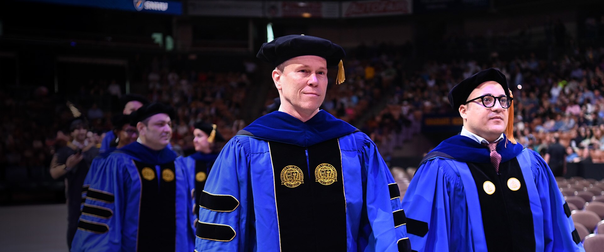 Two male PhD faculty wearing blue, black and yellow graduation cap and gowns at an SNHU Commencement ceremony.