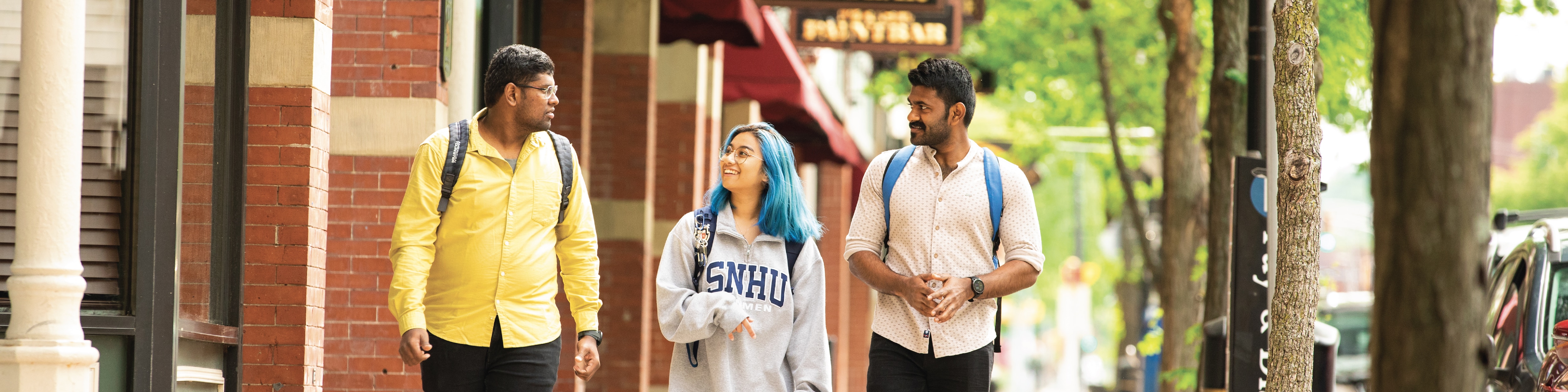 Two young men with dark hair walk alongside a young woman with blue hair in a downtown setting, smiling and engaged in conversation.