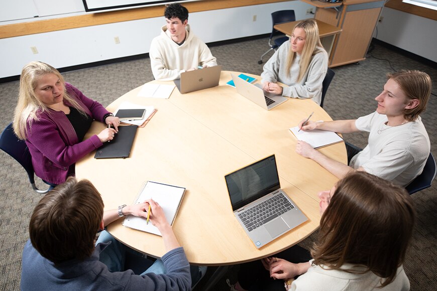 Five students and one instructor sitting around a round table as the instructor speaks to the group.