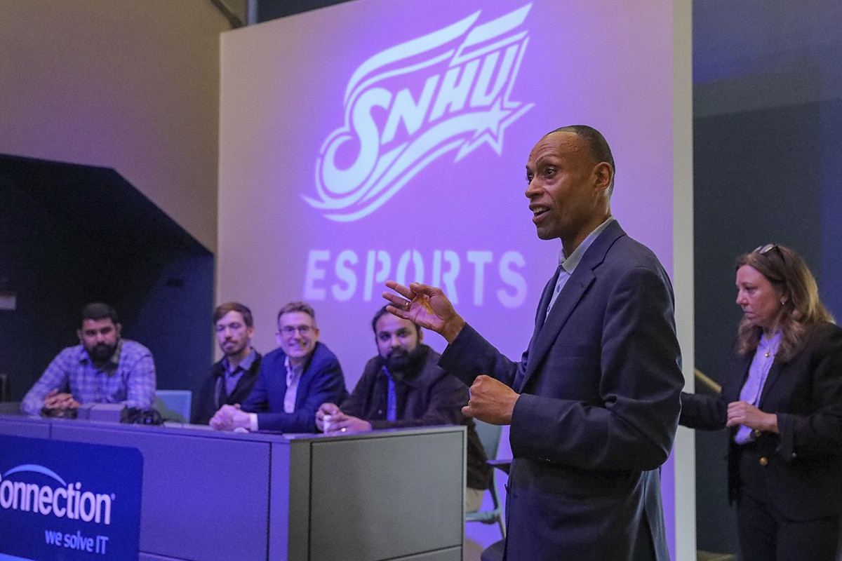 A man, wearing a dark suit, speaking at SNHU's Esports Career Day with attendees in the background. 